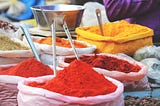 White sacks of spices at an Indian spice market. Each contains an instrument to use to get the spice out of. The image resembles a painter’s palette — mainly different variations of red.