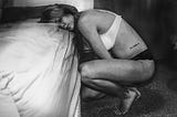 A black and white photo of a woman crouched on the balls of her feet with her head resting on a bed with a look of despair on her face. She is wearing a white bra and black panties. This photo elicits the sense of utter despair that addiction has afflicted the author.
