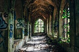 A crumbling stone corridor with arched windows. There is graffiti on some of the walls and ivy breaking through the windows on the right.