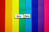 [image description: vertical strips of paper that form a rainbow and a slip of white paper that says “they/them” in black.]