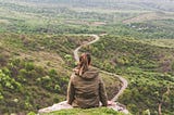 Woman sitting on a hill looking over a long winding road.