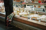 A person in a black outfit stands in front of a brightly lit deli counter, which holds cheeses, and several white bowls of various deli foods.