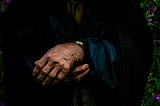 Close-up of an elderly person’s folded hands, worn with age, resting gently against a dark coat — evoking memory, time, and loss.