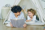 An adult reading and laughing with a small child in an indoor tent
