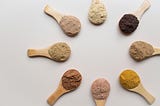 Eight small wooden spoons, each filled with various colors of protein powder, face with their heads inward to form a circle.