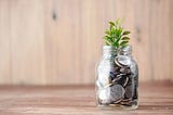 A small green plant growing out of a mason jar will with coins