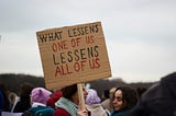 Woman holding a sign that says “What Lessens One of Us Lessens All of Us”
