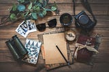 A camera, journals, pencil, glasses and binoculars sit with other items on a wooden surface.