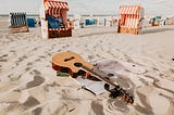 An acoustic guitar, on the beach. A guitar lays upon the sand of a beach. Guitar. A photo of a guitar, alone, laid out on sand. On the beach.