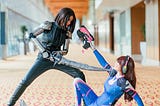 Two women wearing cosplay fighting — one stood up with a sword pointed at the face of the other, who is sat on the ground holding a gun to the first woman’s head