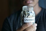 A man, whose face you cannot see, holding a small glass jar full of coins, with the word savings on it.