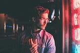 A man in front of some neon lights, holding his own hand.