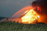 Firefighter aims water hose at burning wood shed in a farm field.