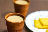 Two clay cups filled with steaming Indian tea (chai) placed on a wooden table, accompanied by a plate of crispy snacks in the background.