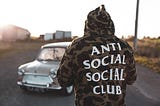Man wearing hoodie that says anti social social club, standing in front of a car