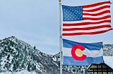 A photo of the American flag and Colorado flag with snow-covered mountains in the background taken by dannyboy4125 via Unsplash.com