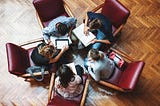 Five people sit in a circle on red office chairs. They are huddled closely together with notebooks on their laps, appearing to be discussing and working together.