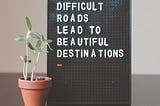 Little green plant next to black and white sign which reads “Difficult roads lead to beautiful destinations”