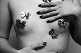 A naked chest of a person with butterfly pasties, and their hands with painted nails touching their body.