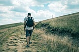 Man in white tee shirt and black shorts carrying a black back pack walking up a dry, barren hill toward a cross on a hill. The author is using the picture to show that having faith in the way to get somewhere is not always clear, but to follow God’s guidance and you will arrive.