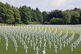 green field with dozens of white crosses in ground with green forest in background, symbolizing graves of soldiers lost to war posted on mickey markoff 2024 gold star mothers and families day article