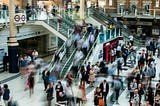 Wide angle view of a train station with multiple escalators and sets of stairs and many people. There are some shop fronts visible in the picture. Some of the people are blurry with movement.