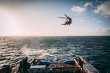 A helicopter flys above the ocean following behind the stern of a vessel