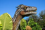 A dinosaur’s neck and head in profile sticks out of vegetation. He has big teeth and his green beady eye is looking up at the sky