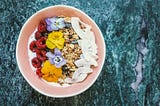 A bowl of vegan yoghurt topped with fresh raspberries, edible flowers, granola, seeds and coconut flakes, arranged on a green‑blue marble surface.
