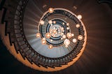 Looking up at a spiral staircase with many light bulbs hanging in the middle.