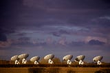 A row of radio telescope dishes aligned in the same direction across a flat landscape, lit warmly against a dramatic cloudy sky at dusk.