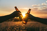 Two disembodied arms each hold a bottle of beer, toasting each other in front of a setting sun, such that the sun gleams through the brown bottles. (Though I suppose it could also be a sunrise, in which these two are some seriously committed day drinkers!)