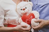 A couple standing close together, holding a white teddy bear with a red heart that reads “love,” their hands gently touching around the plush toy.