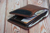 An image of a brown leather wallet with black trim on a rustic table. It has quite a few bills, cards and receipts in it — but it’s nowhere near as stuffed as mine is!