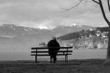 Old man alone on a park bench beside a lake with mountains in the background