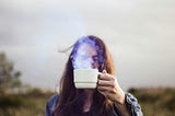 A person with long brown hair holds a cup of tea up to the camera, their face mysteriously obscured by the cup itself and the steam rising from it. The background is an out-of-focus rural scene.