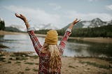 A woman in flannel looks over a mountain landscape with her arms in the air.