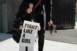 A woman with a little girl. The little girl is holding a sign that reads, Fight Like a Girl.