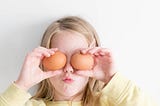 A young girl holding fresh eggs over her eyes — Not unlike my first baking experience!