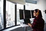 A woman with orange headphones and glasses sitting in front of two monitors and a laptop looking out the window in a metropolitan environment.