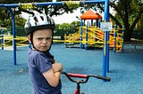 Photo shows young boy with bike and helmet upset at playground closure. Boy has arms crossed and scowls toward camera as yellow caution tape is wrapped around playground equipment.