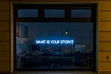 A neon sign over a desk reads, “What is your story?”