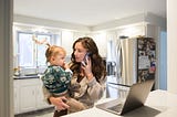 anxious mom on phone while holding toddler in front of open laptop caitlin houston 