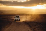 A vehicle driving down a dirt road. Dust drifts away from it as it heads towards the sunset.