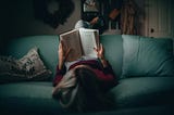 A woman lays on a blue-green sofa reading a book, so that we can see the pages.