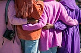The backs of four Black women in brightly colored coats. They each have their arms around one another.