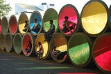 6 people casually sitting inside large cement pipes stacked in two rows, and covered on one opening side with blue, green, red, yellow film — to let filtered coloured light pass