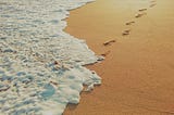 Surf foams washing over a set of footprints on a sandy beach.