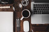 A laptop on a table accompanied by a cup of coffee, two pens, a spiral notebook and a magnifying glass.