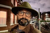 Selfie of brown man with glasses in green felt fedora and wax cotton jacket. Street with houses in the background. Cloudy dusk sky. Street lighting.